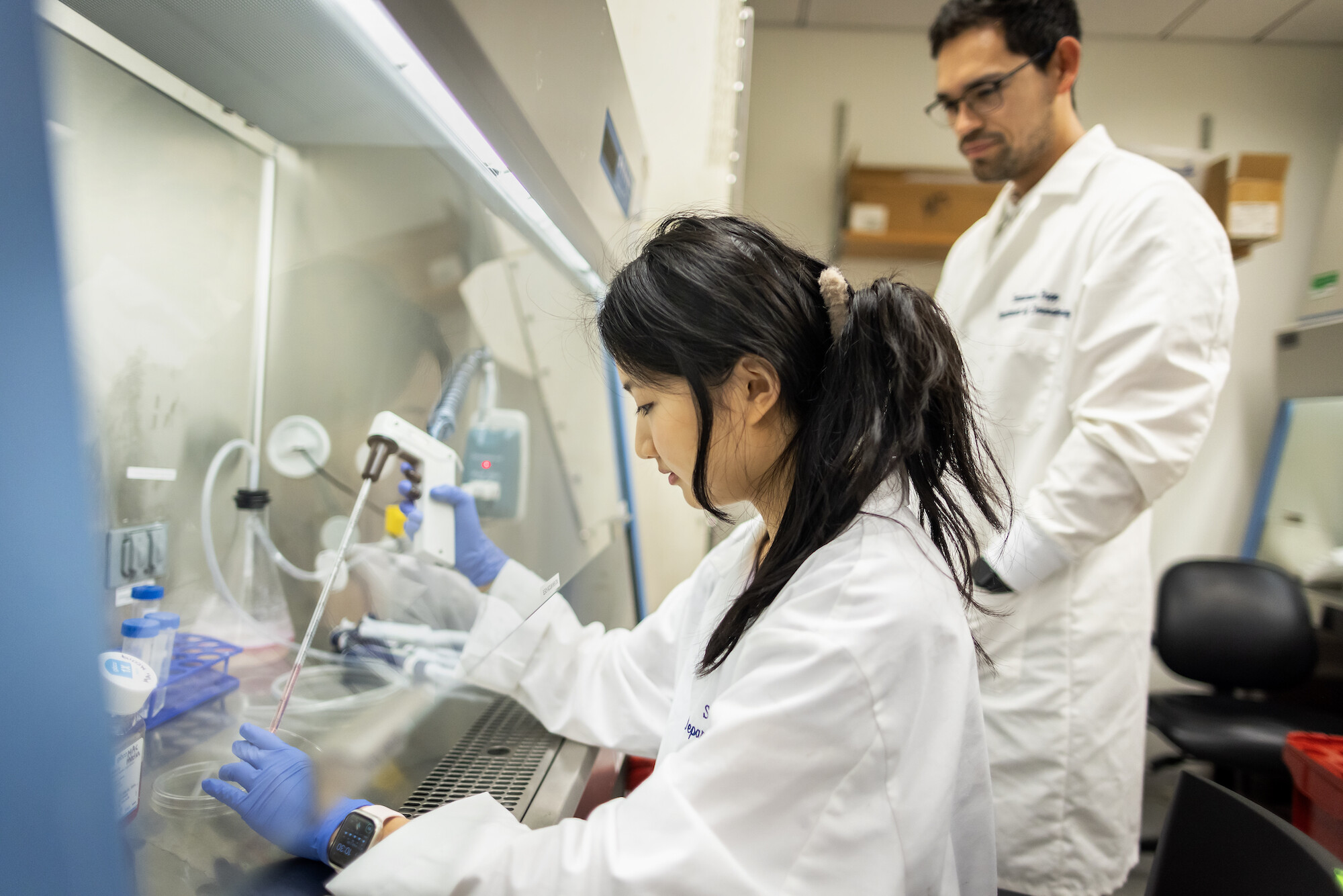 Jiayi Pang, seated wearing a white lab coat, uses a pipette to fill a test tube as her mentor looks on