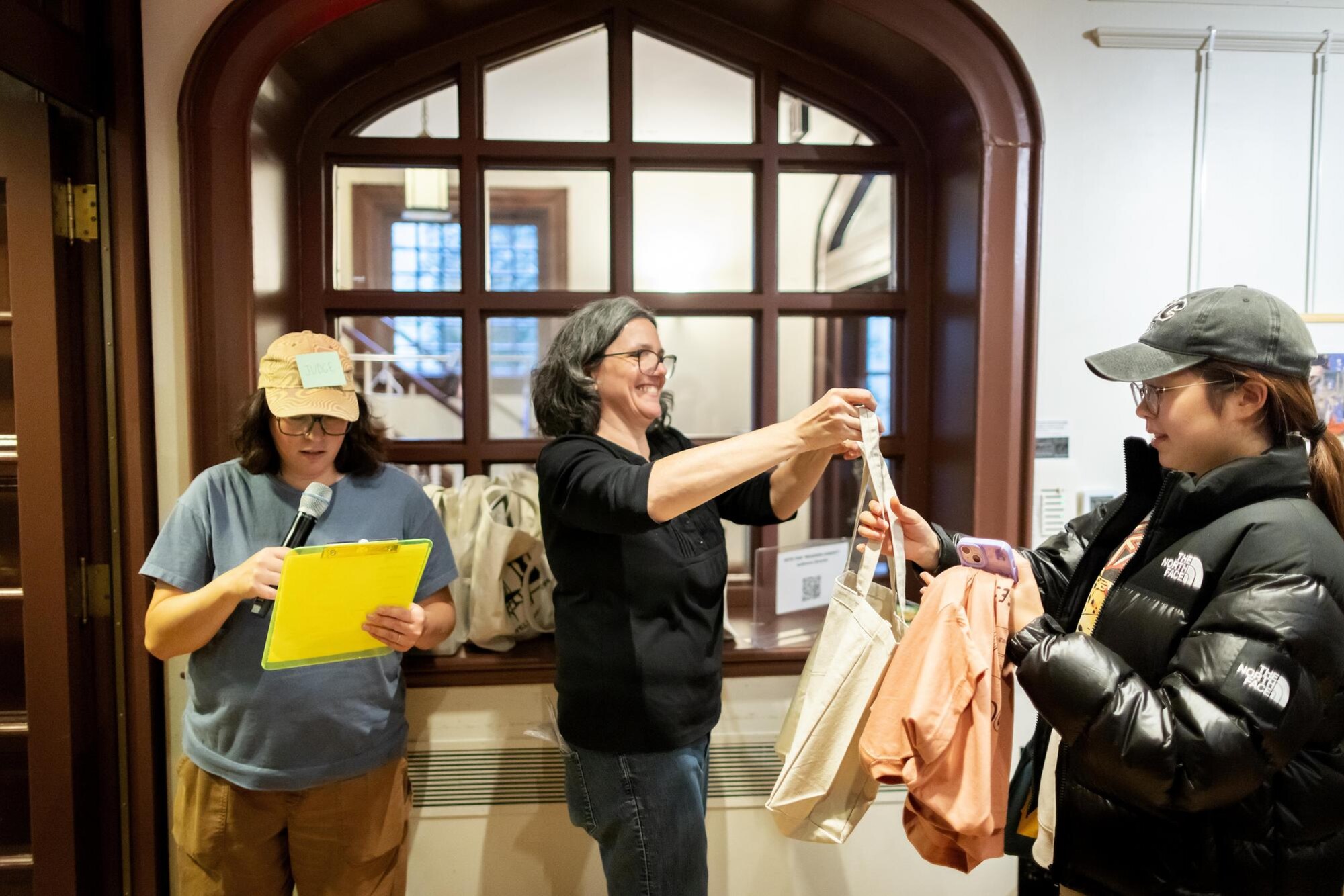 A person hands another person a tote bag next to a person with a clipboard and a microphone.