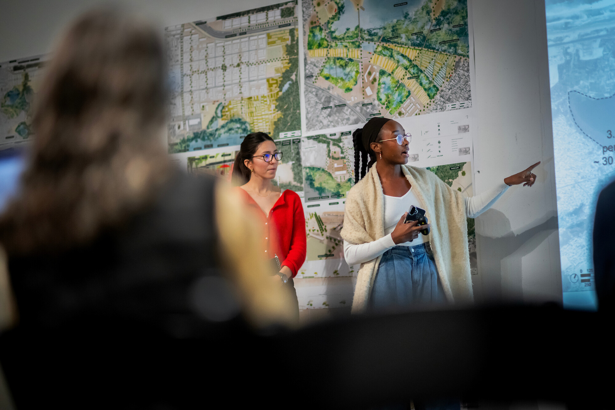 A presenter gestures towards a projected map while another person stands beside her.