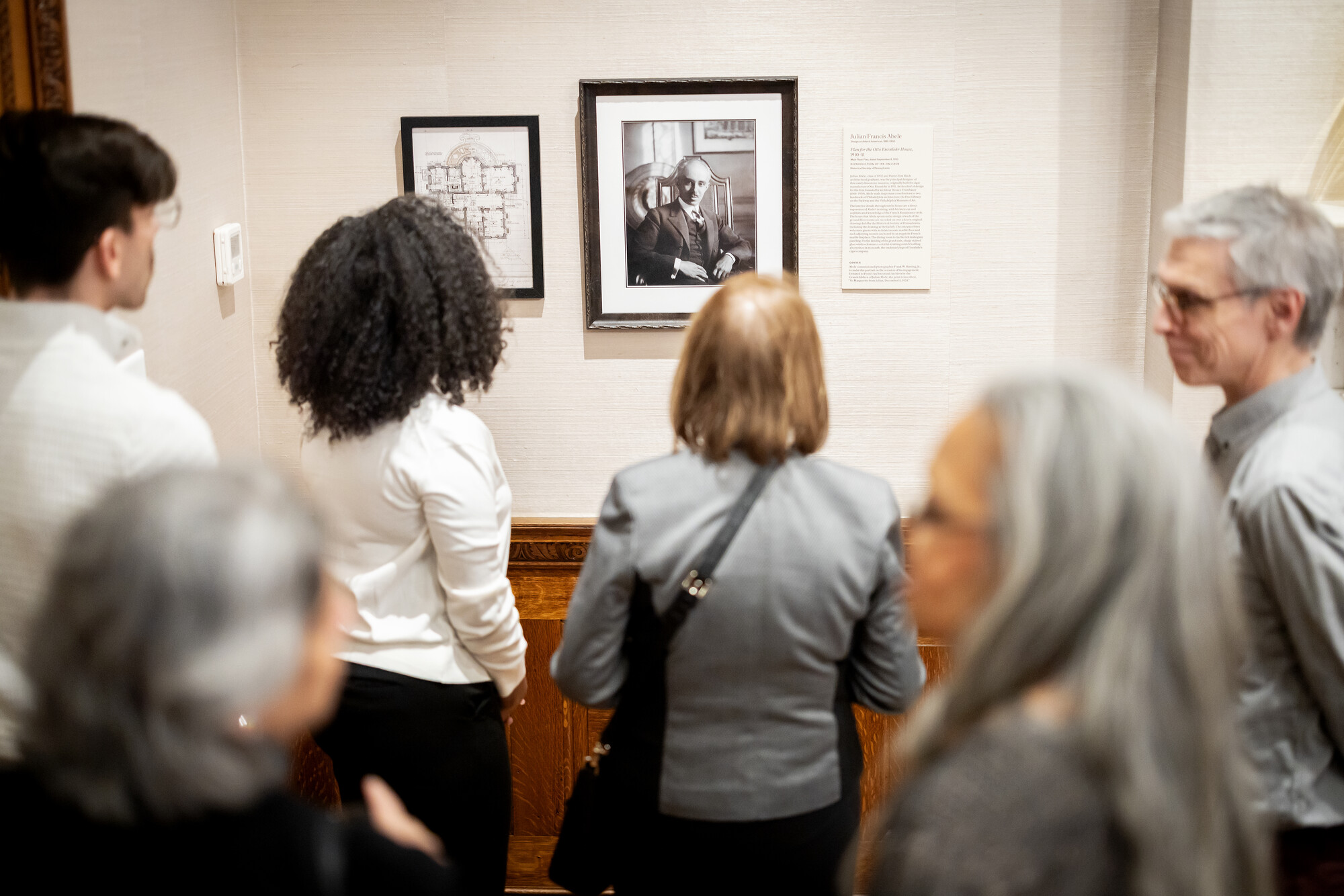 Crowd gathers around a framed portrait and floor plan.