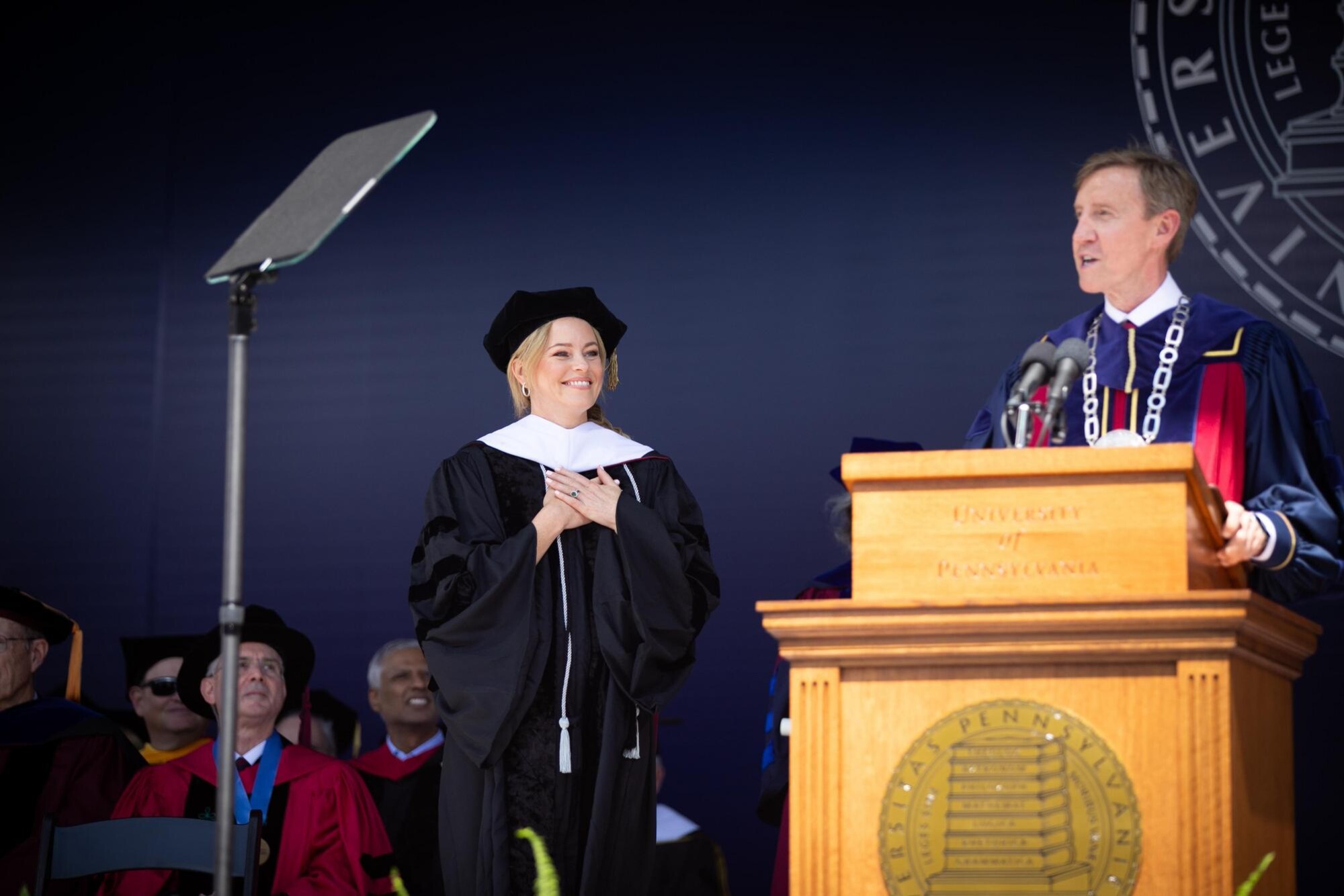 Elizabeth Banks on the stage as J. Larry Jameson delivers a speech at the podium.