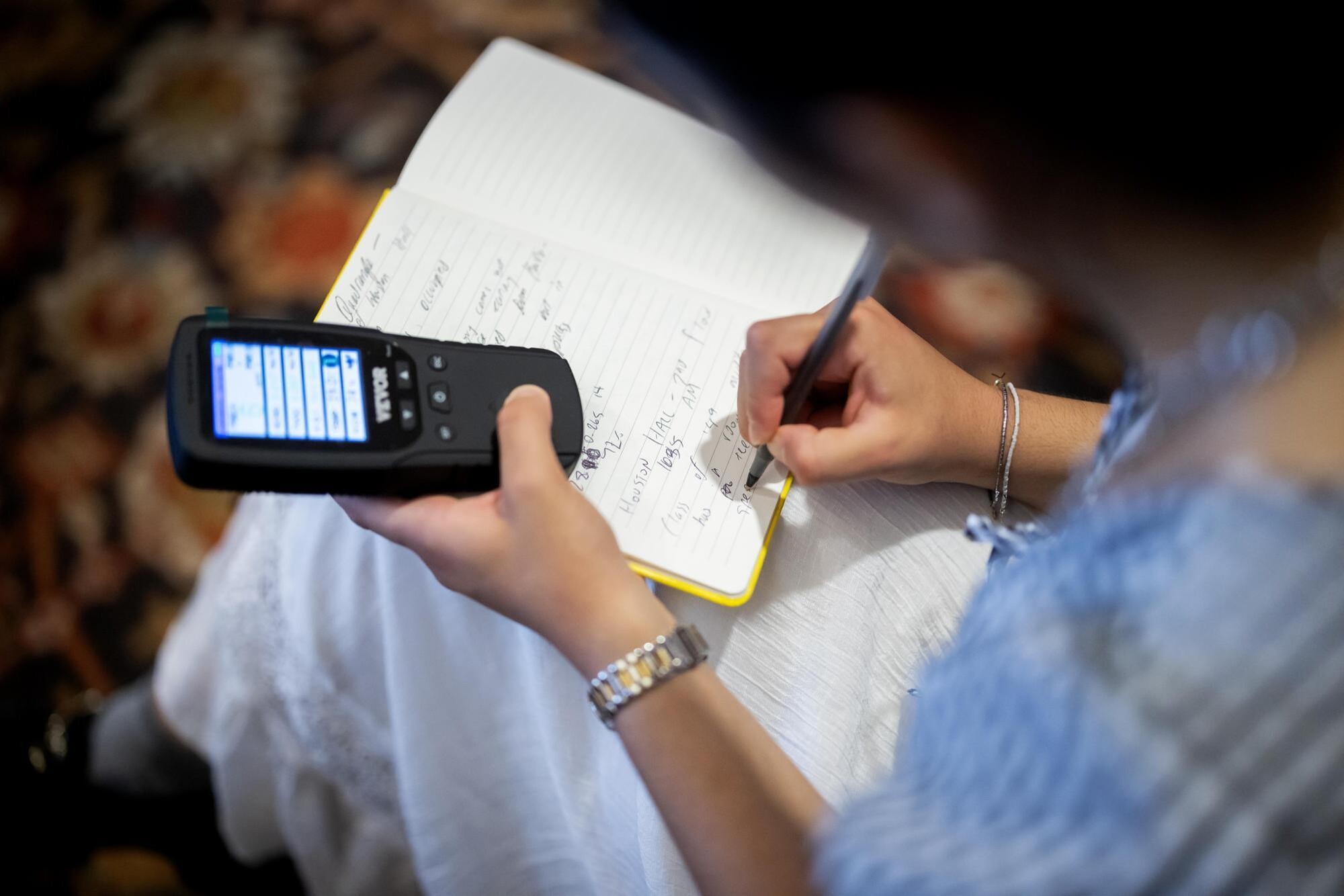 A student making notes in a notebook while holding a digital device.