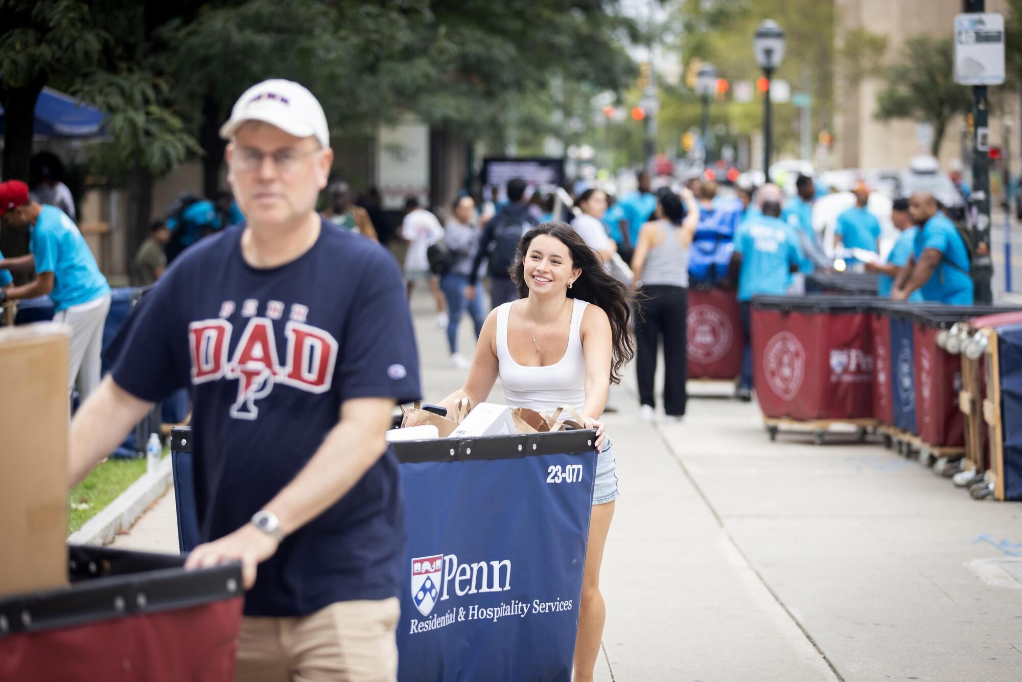 A Penn student and a man wearing a Penn Dad T-shirt push Move-In carts on a sidewalk during Move-In.
