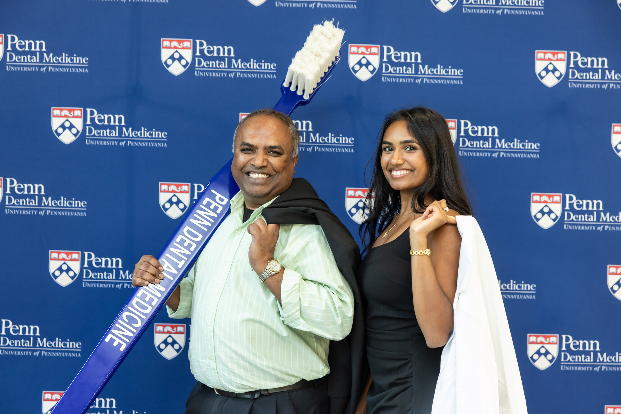 Students posing for a photo at a Penn Dental step and repeat, one holding a white coat the other holding a large prop toothbrush.