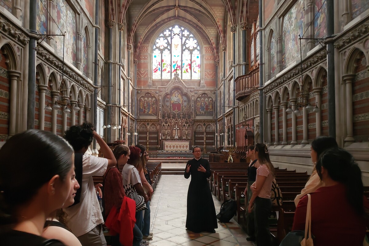An ornate Anglican church with stained glass. Students stand near the pews listening to a frocked speaker.