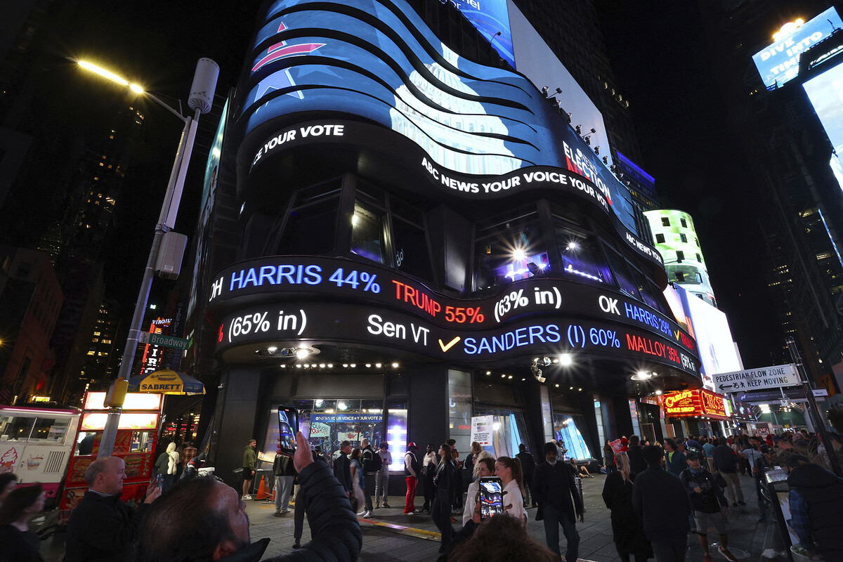 A side-scrolling election feed is displayed on the side of a building in Times Square in New York City. There are people below in the foreground.