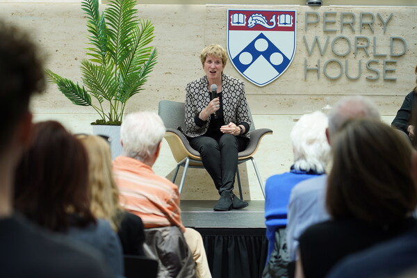 Kate Gilmore sits on a chair on a stage holding a microphone in front of audience members and a Penn shield and the words Perry World House are behind her.