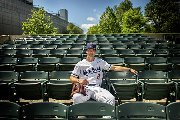 Wyatt Henseler sits in the bleachers at Meiklejohn Stadium while wearing his white jersey and his right arm resting on a baseball glove.