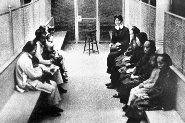 A group of Chinese and Japanese women and children waiting to be processed, held in a wire mesh enclosure. Benches line either sides of the room, with a stool in the middle.