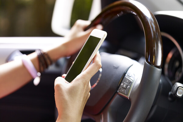 A teenagers hands holding a steering wheel in one hand and a smartphone in the other.