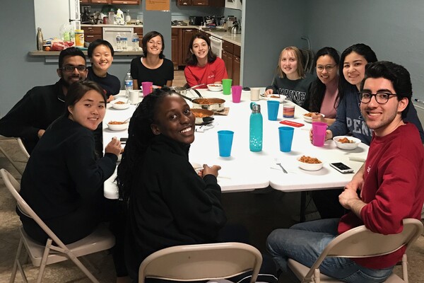 Ten students around a table all looking and smiling at the camera. The table has bowls of food and cups and is next to a kitchen. 