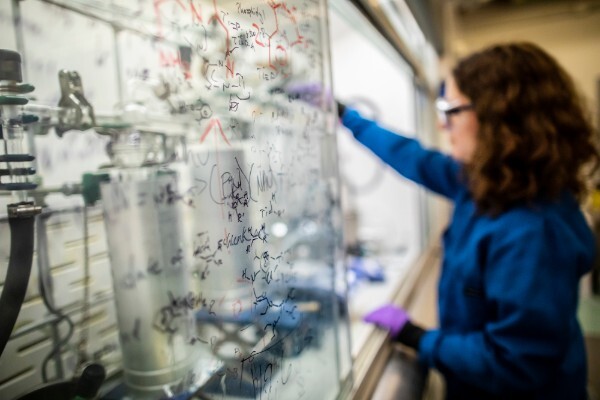 a close-up of chemical reactions written on a glass screen with a person in a lab coat working in the background