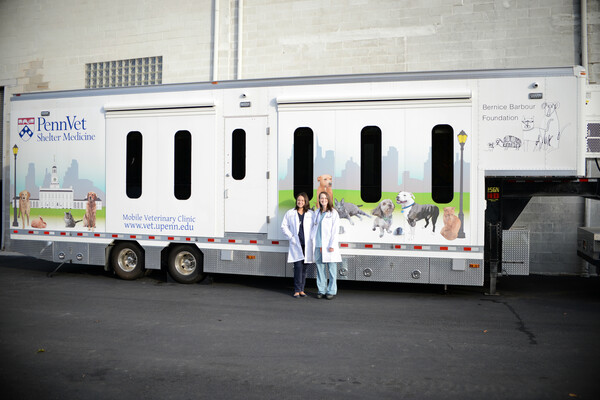 Two women stand in front of a trailer with the words "Penn Vet Shelter Medicine" emblazoned on it and pictures of dogs and cats on the front 