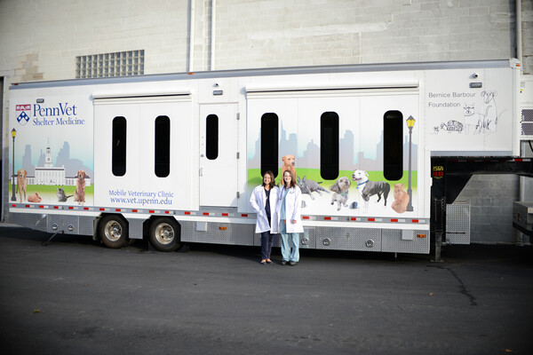 Two women stand in front of a trailer with the words "Penn Vet Shelter Medicine" emblazoned on it and pictures of dogs and cats on the front