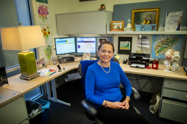 Woman in blue shirt sitting with desk behind her. Desk is filled with lamp, two computer screens, a printer and a coffee mug tree.