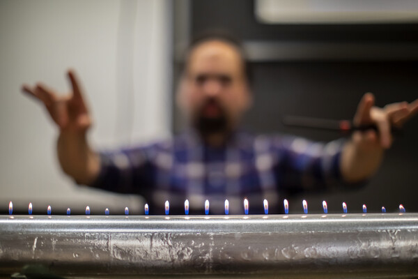 a metal tube with a line of flames coming out of the top and a blurred person posing in the background