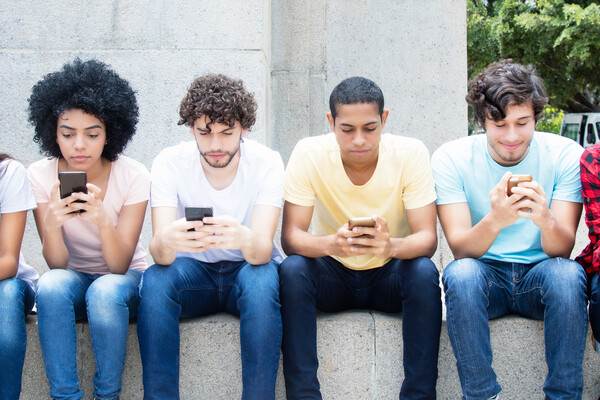 Teenagers of various races and genders sitting on a stone wall with cell phones in hand. 