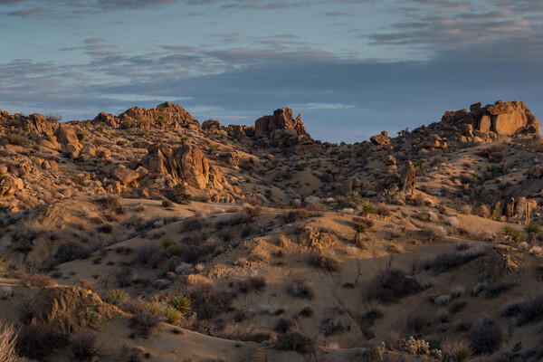 A rocky, shrubby landscape glows with sunlight under a partially cloudy sky.