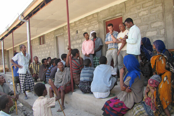 A few people stand in front of a building talking to a larger group of gathered people listening.