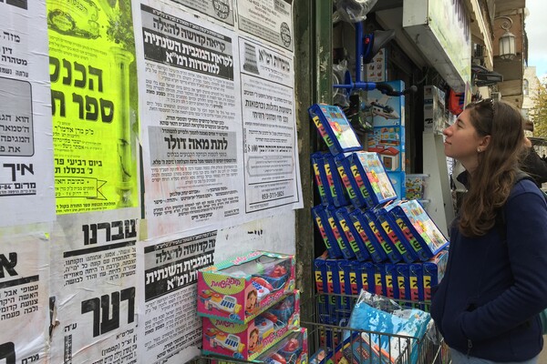 A girl in a blue fleece standing in front of a wall of black and white posters in Hebrew. Boxes of children's toys are in front of the wall.