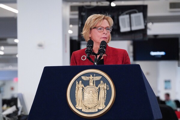Woman in a red jacket standing at a podium with two microphones.