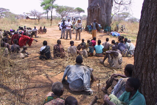 A large group of people sit on the ground outside, roughly in a circle around a group of presenters in the African landscape.