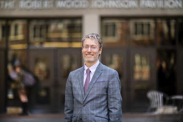 A man in a blue plaid coat, pink shirt and purple tie standing in front of a blurry building.