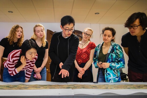 Seven students gathered together looking at a painted Chinese scroll unrolled on a table.