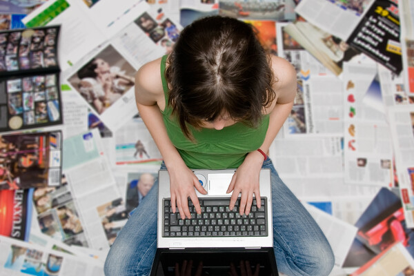 a person sitting on a stack of open magazines and newspapers working on a laptop