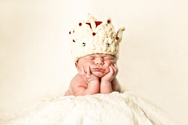 Newborn on a blanket wearing a crown resting their head on their hands.