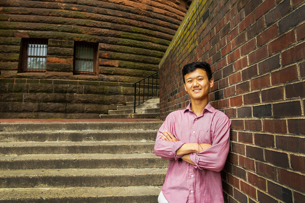 A person in a pink button-down shirt leaning against a brick wall. 