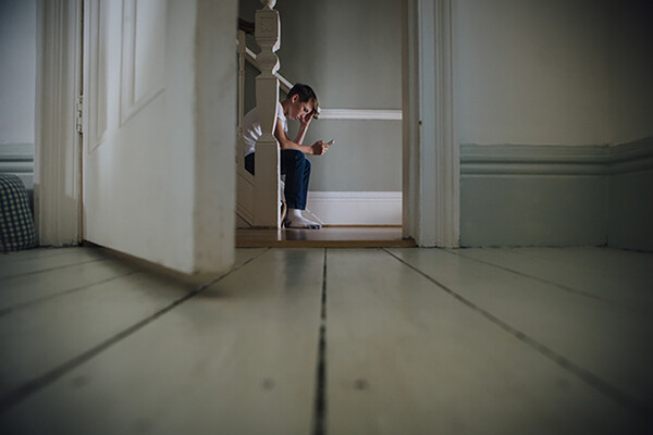 open door with young person sitting on bottom stair of a staircase in background, looking down at a phone in hand.
