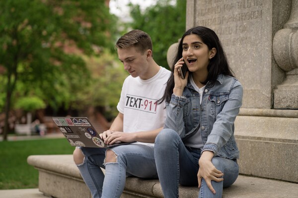 Two students sitting on a stone statue, one on a computer, the other on a phone.