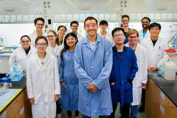 a group of people standing in a chemistry lab wearing lab coats and goggles