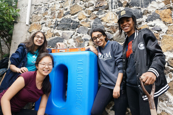 Four smiling students posing by a blue container with a level on top of it