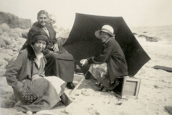 Gladys Tantaquidgeon seated on a beach with four other people and a black umbrella on the ground.