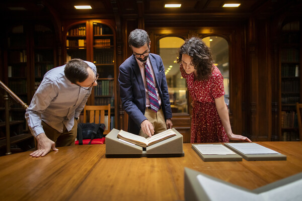 Three people standing over a book in a library setting. 