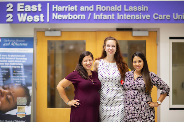 A tall red-headed smiling young woman stands between two pregnant smiling women, under a sign that says 2 East 2 West Harriet and Ronald Lassin Newborn/Infant Intensive Care Uni