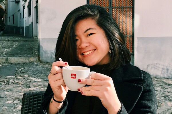 Student sitting outside holding a coffee cup and smiling. 