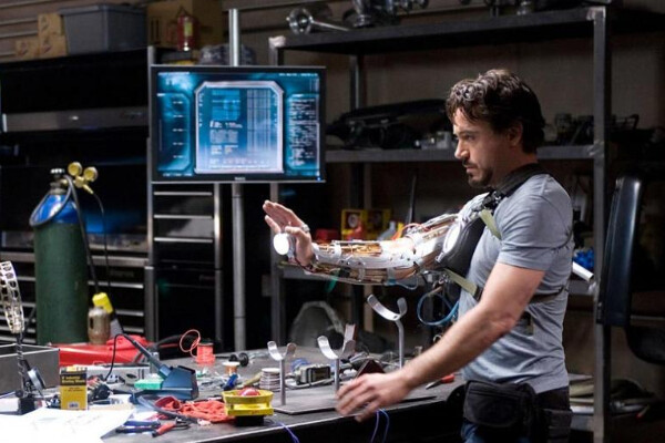 Film still of Robert Downey Jr.'s Iron Man character standing at a work table full of tools trying on a robotic-looking arm.