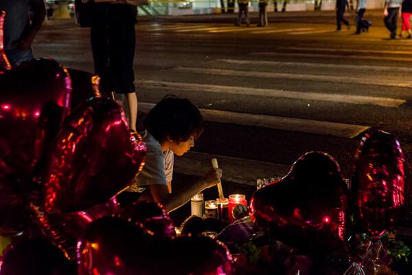 young child kneeling on ground lighting a candle surrounded by heart balloons at night, at a vigil for a mass shooting.