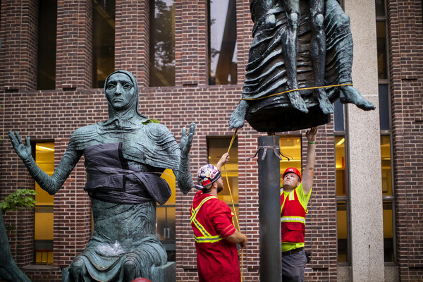 A sculpture of a female figure and the feet of another sculpture of two figures as they are being lowered onto a pipe while one construction worker holds a rope and another reaches up.