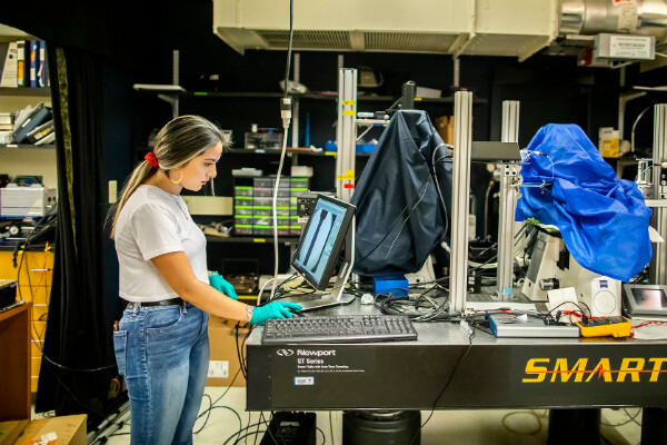 angelica padilla working in a lab on a crowded optics table looking at a computer