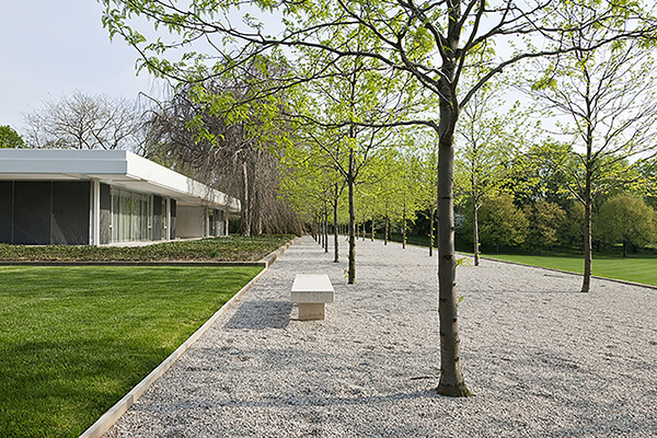 Exterior view of Miller House, with a pebble courtyard with trees and a bench, green lawns and a modernist home.