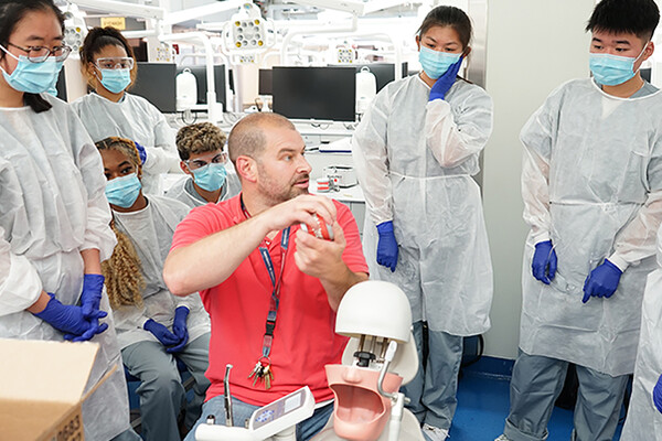 A Penn Dental Medicine instructor sits with a dental model tool in hand beside a replica of head with an open mouth while seven high school students in white lab coats, gloves and masks watch the demonstration.