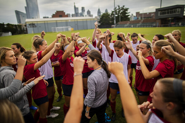 Players on the women's soccer team gather in a circle with their fists raised with their coach at practice.