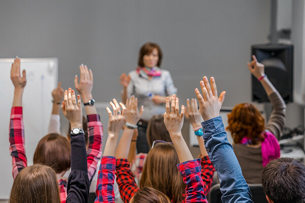 Seated students in a classroom raise their hands while a teacher stands at the head of the classroom