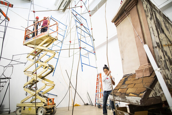 Artist with hands on long metal pipe with fragment of a building behind her and scaffolding hanging from the ceiling during sculpture installation.