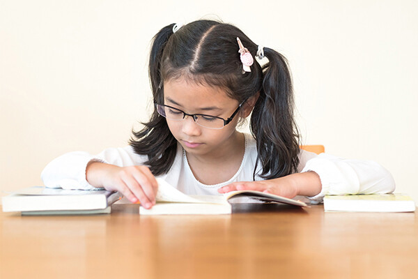 A child sits at a table reading a book, two closed books are on the table beside them.