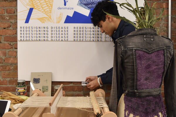 student setting up design exhibit with a jacket and a loom and a sign.